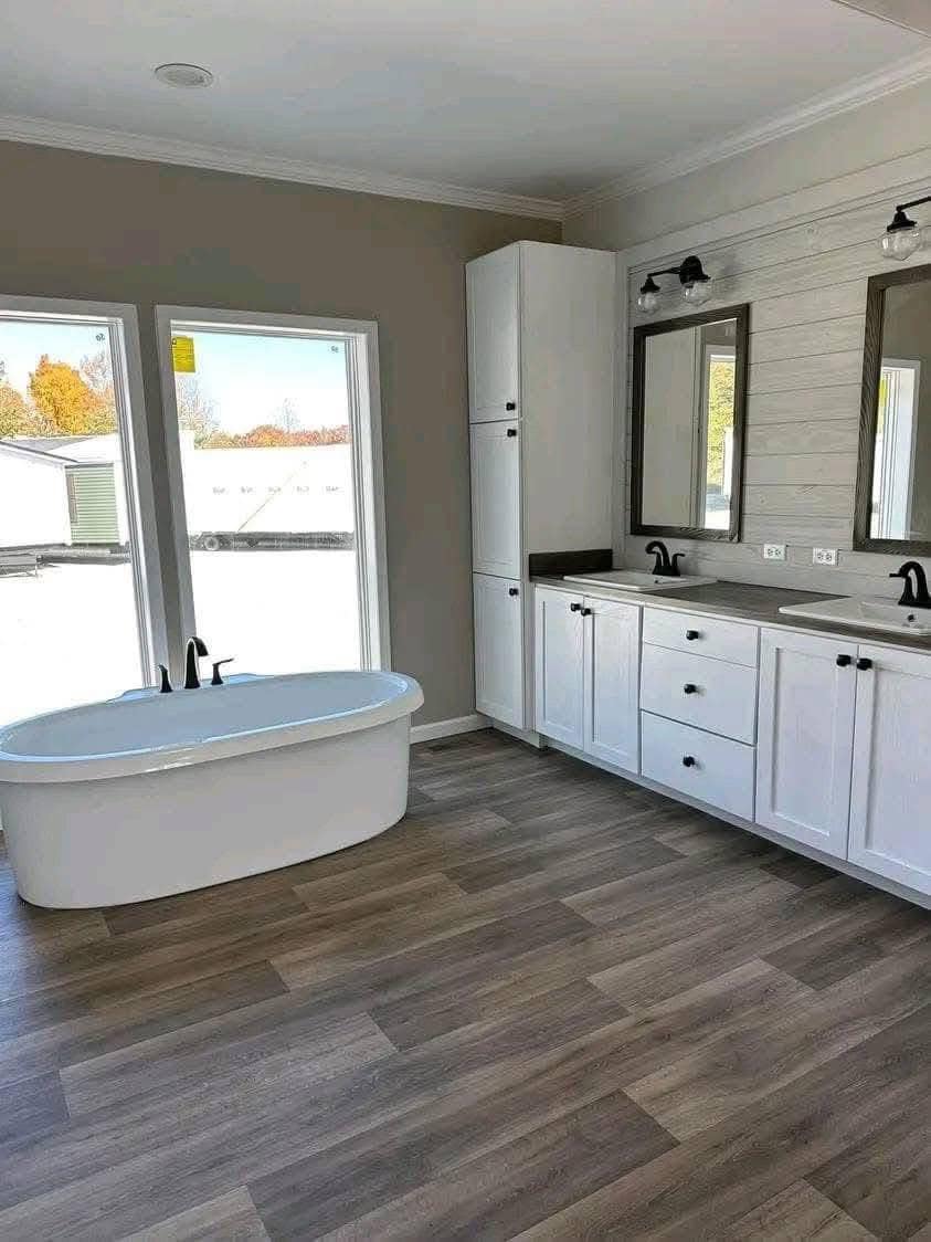 Spacious bathroom with a freestanding white tub by large windows, dual white sink vanity with black fixtures, and wood-look flooring, creating a serene ambiance.