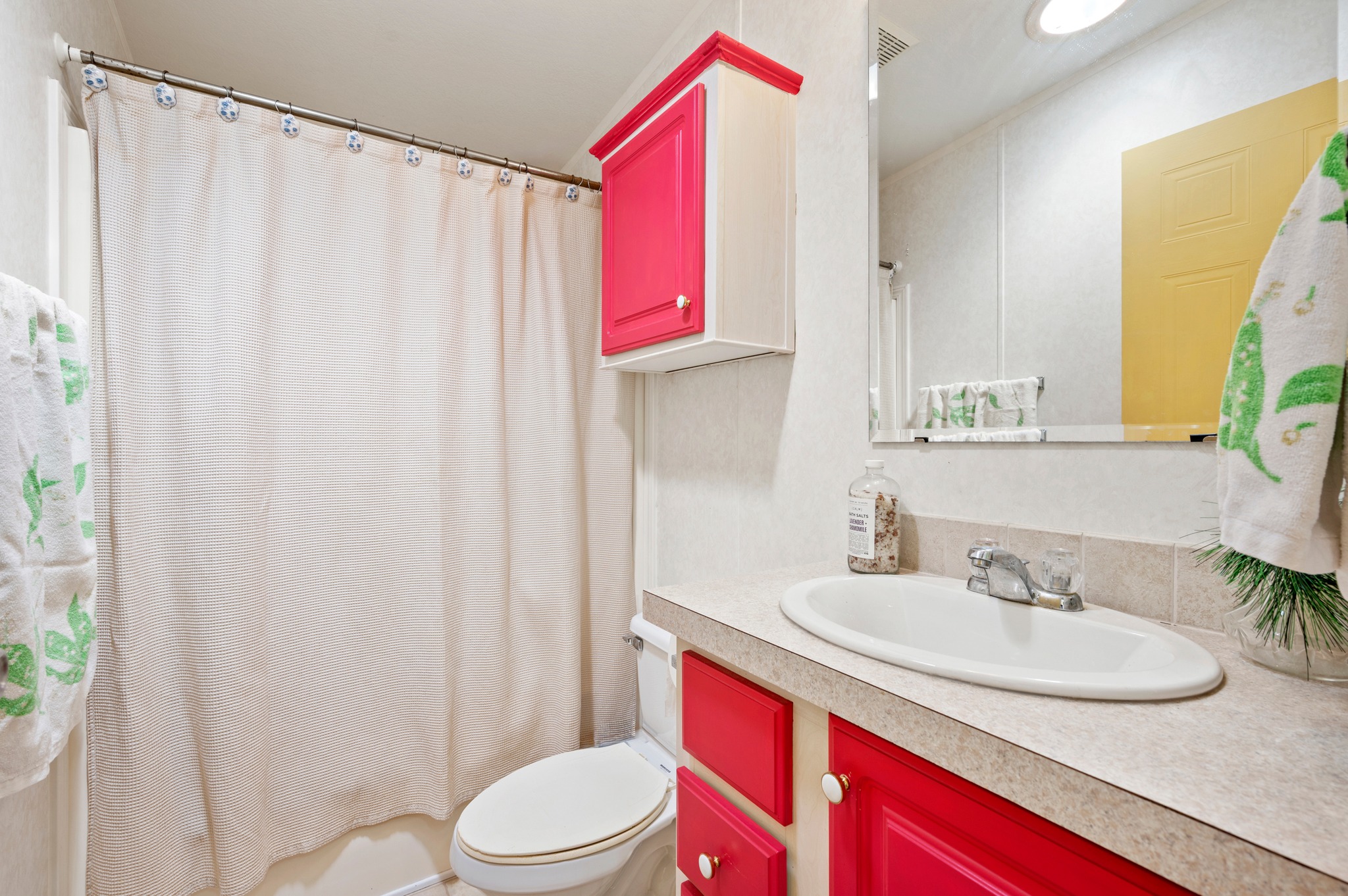 A small bathroom with beige walls features vibrant red cabinets and drawers. A white sink and faucet sit on the light countertop. A beige shower curtain and green-accented towels add a fresh feel.