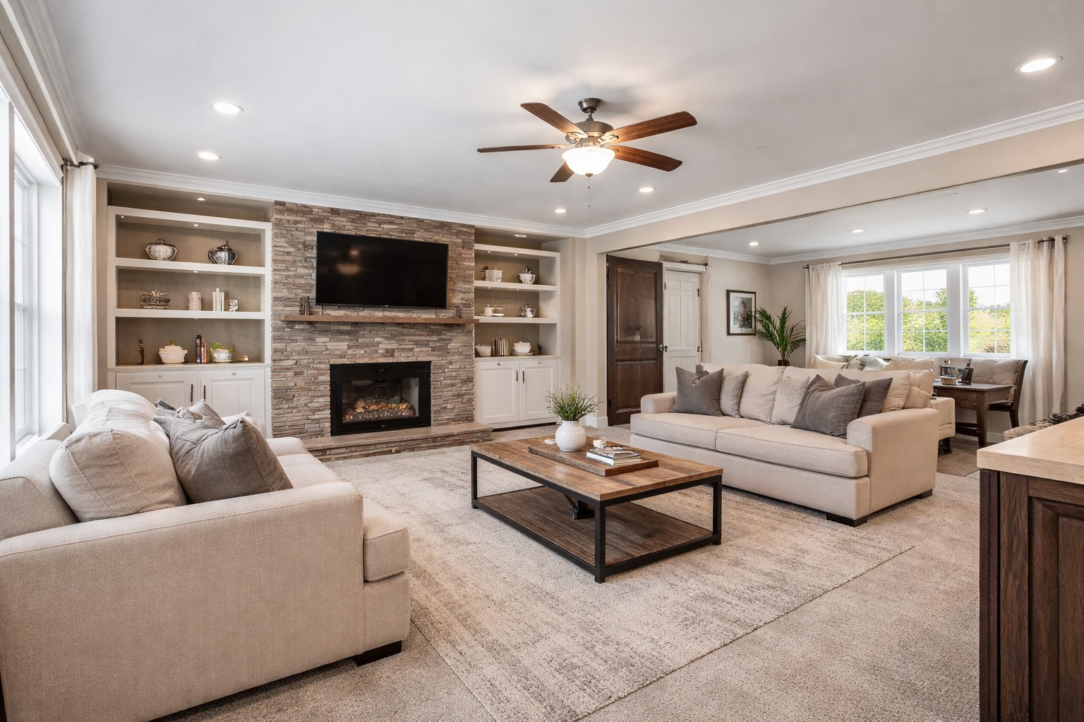Spacious living room with beige sofas, a wooden coffee table, stone fireplace, and built-in shelves. Ceiling fan and large windows provide a cozy feel.