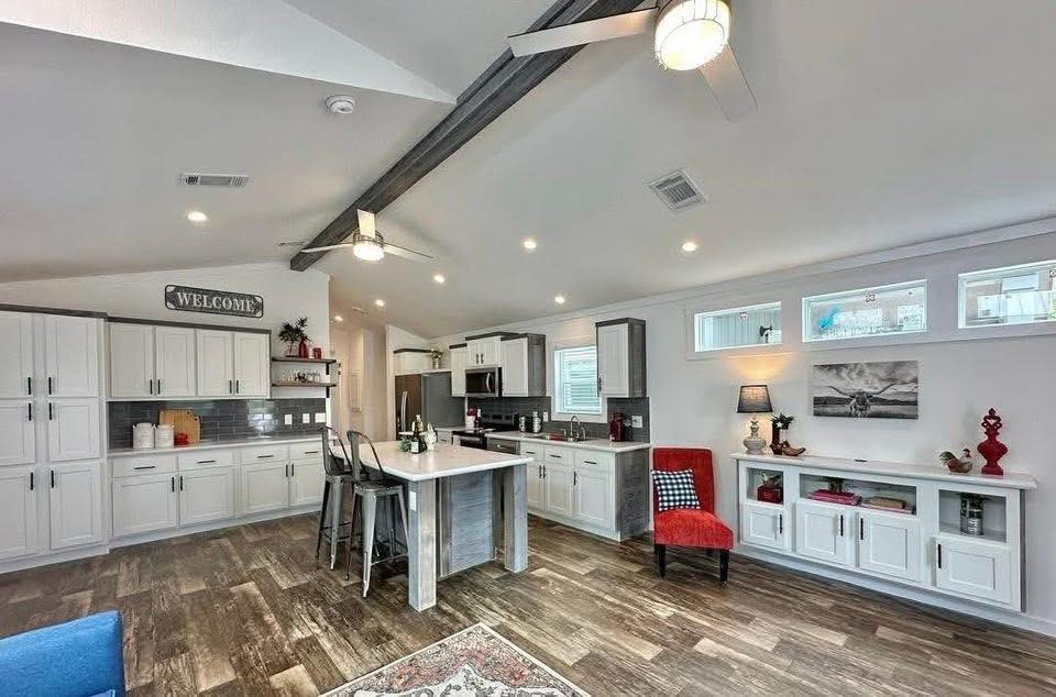 Spacious modern kitchen with white cabinets, a gray island, and wood flooring. Bright lighting, red chair, and decorative elements create a welcoming feel.