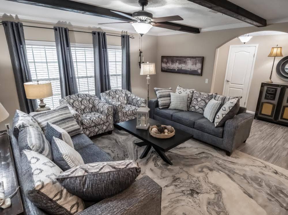 A cozy living room with gray sofas and patterned armchairs placed around a black coffee table. Soft lighting and a large window create a warm, inviting atmosphere.
