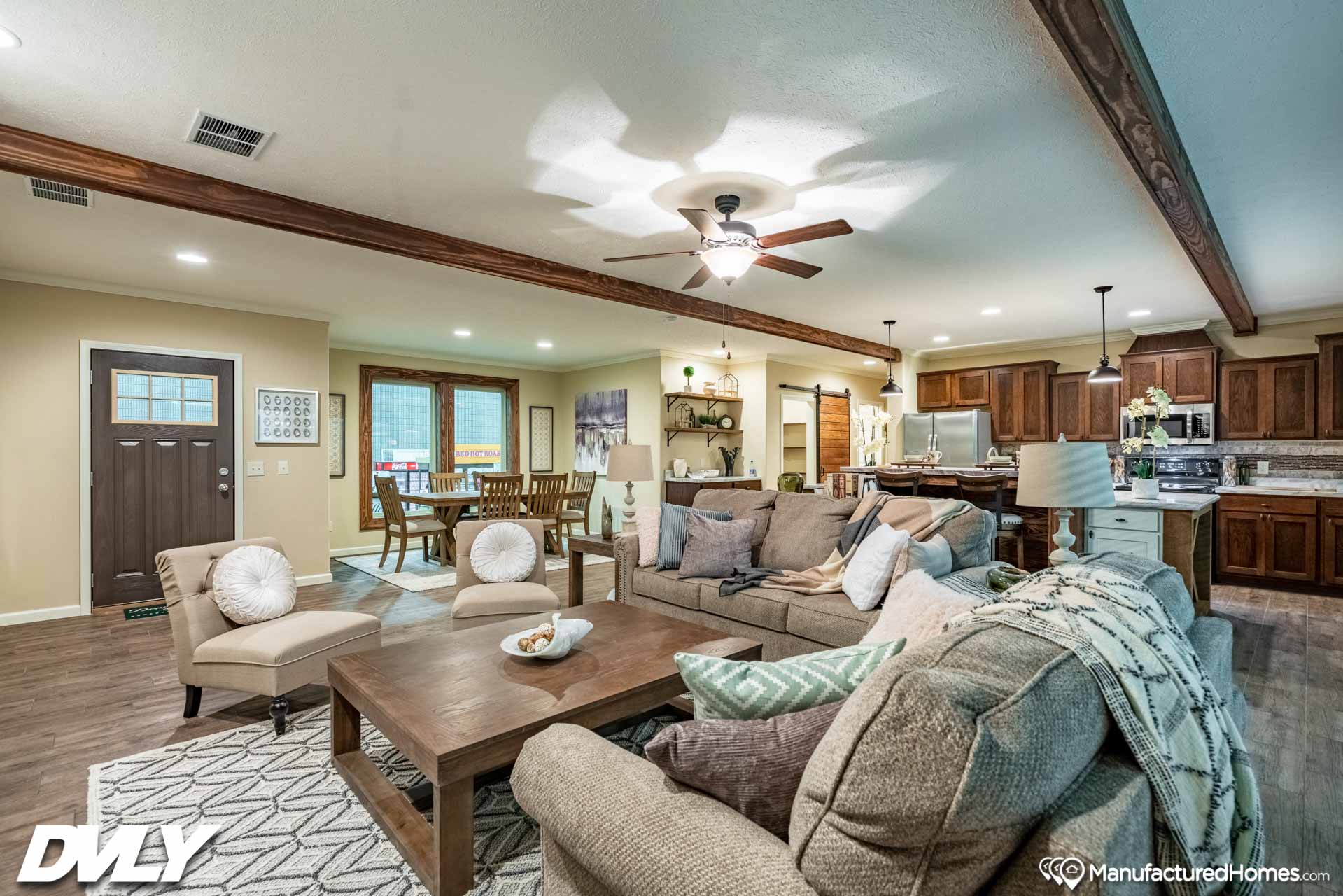 Stylish open-concept living space with cozy sofas, a wooden coffee table, a dining area, and a kitchen. Warm lighting, ceiling fan, and rustic beams create an inviting atmosphere.