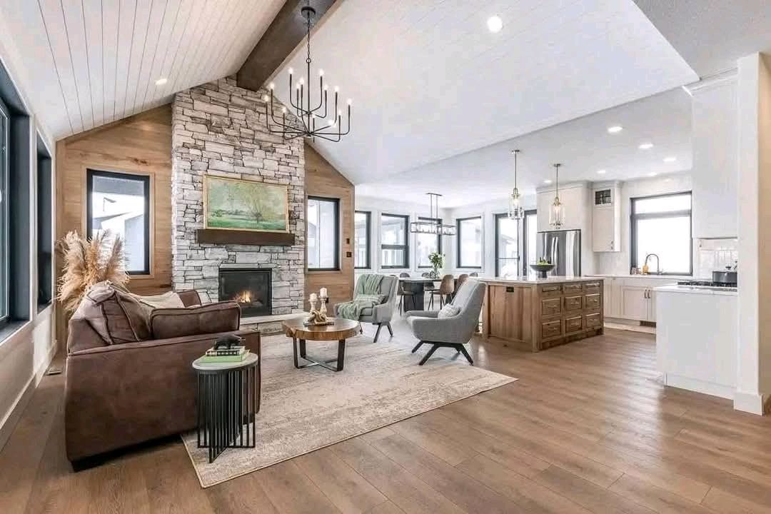Spacious living area with high ceilings, stone fireplace, and large windows. A brown sofa, neutral rugs, dining area, and kitchen create a cozy, modern vibe.