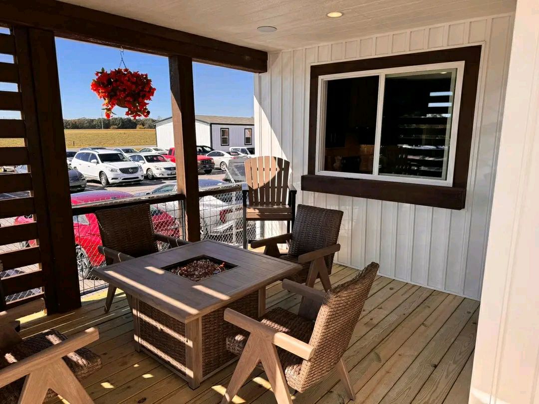 Cozy porch with wicker chairs around a fire pit table on wooden decking. Hanging red flowers and parked cars in the background create a peaceful vibe.