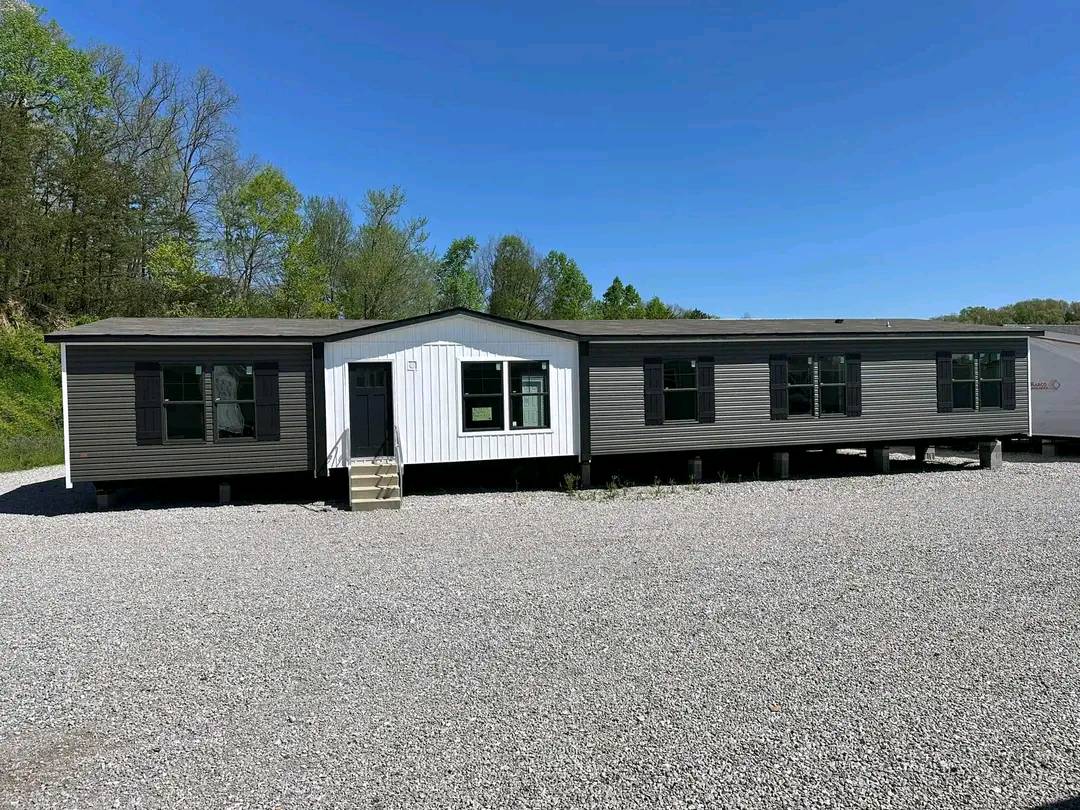 A large modular home with gray siding and black shutters sits on a gravel lot. Surrounded by trees under a clear blue sky, it exudes a peaceful, rural feel.