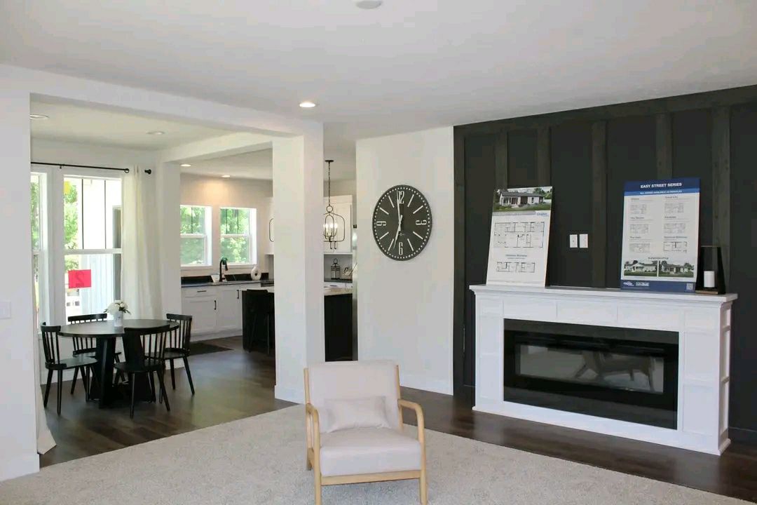 Modern living room with white walls, featuring a large black clock and fireplace. Open layout to kitchen and dining area with dark wood flooring.