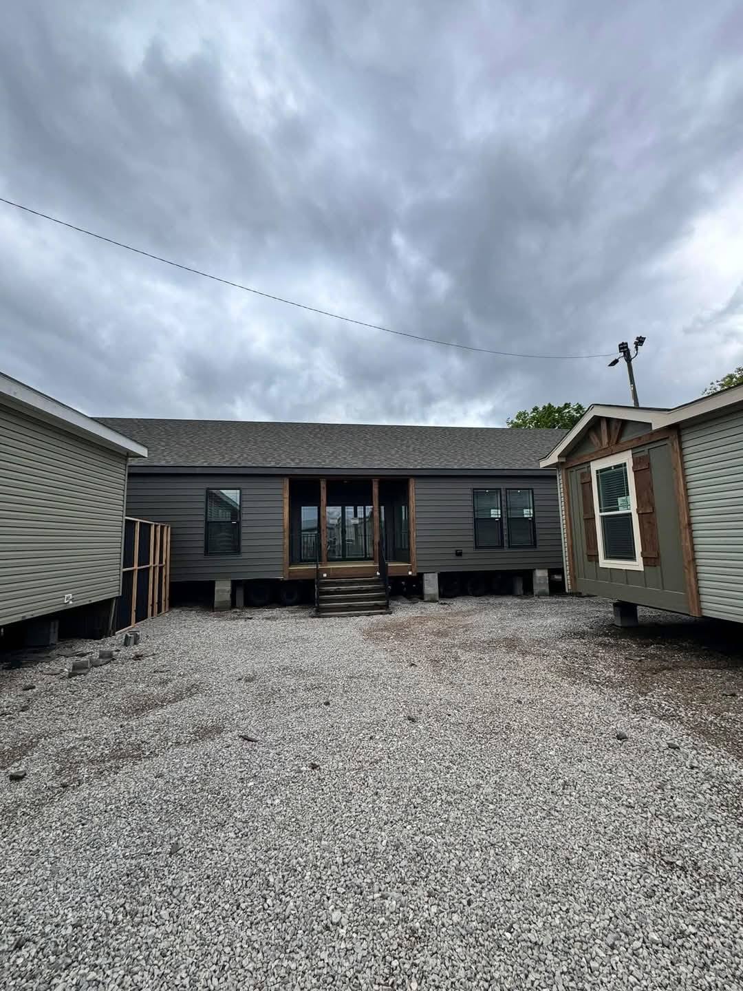 Dark gray mobile home under overcast sky, surrounded by gravel ground. Two other mobile homes are partially visible on each side, creating a quiet, moody atmosphere.