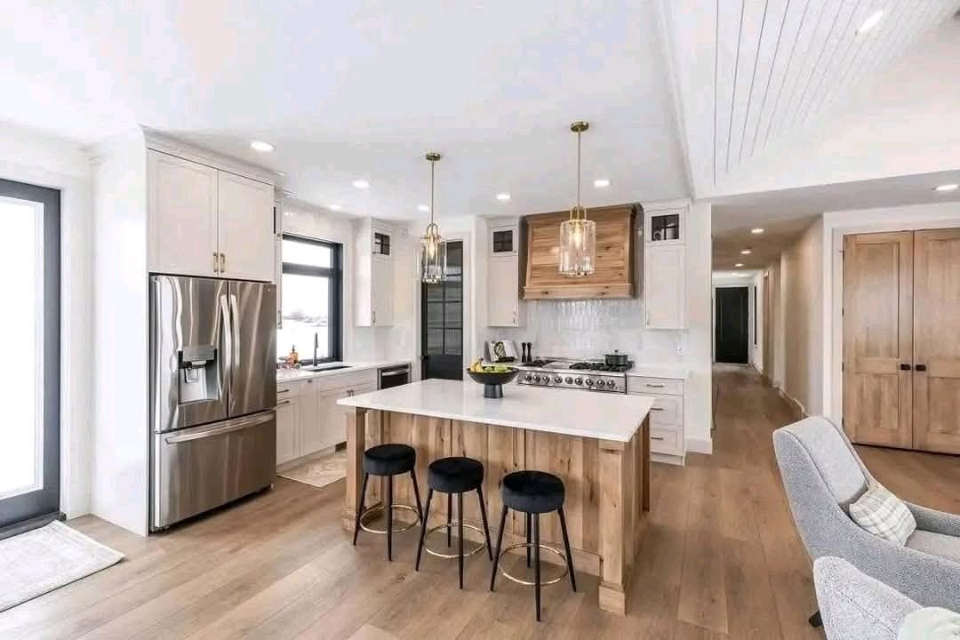 Modern kitchen with white cabinets, stainless steel fridge, and a large island with wooden accents. Three black stools sit by the island. Bright, airy atmosphere.