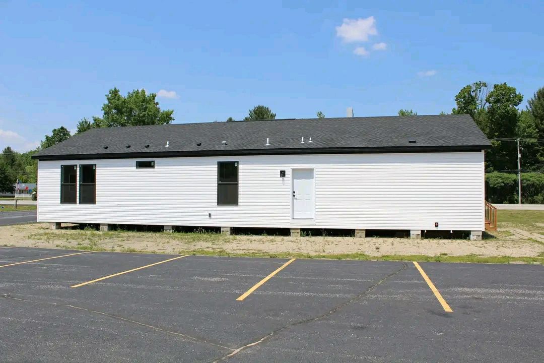 Single-story white modular building with a black roof, set on a sandy lot. Windows and a central door are visible. Empty parking lot in foreground.