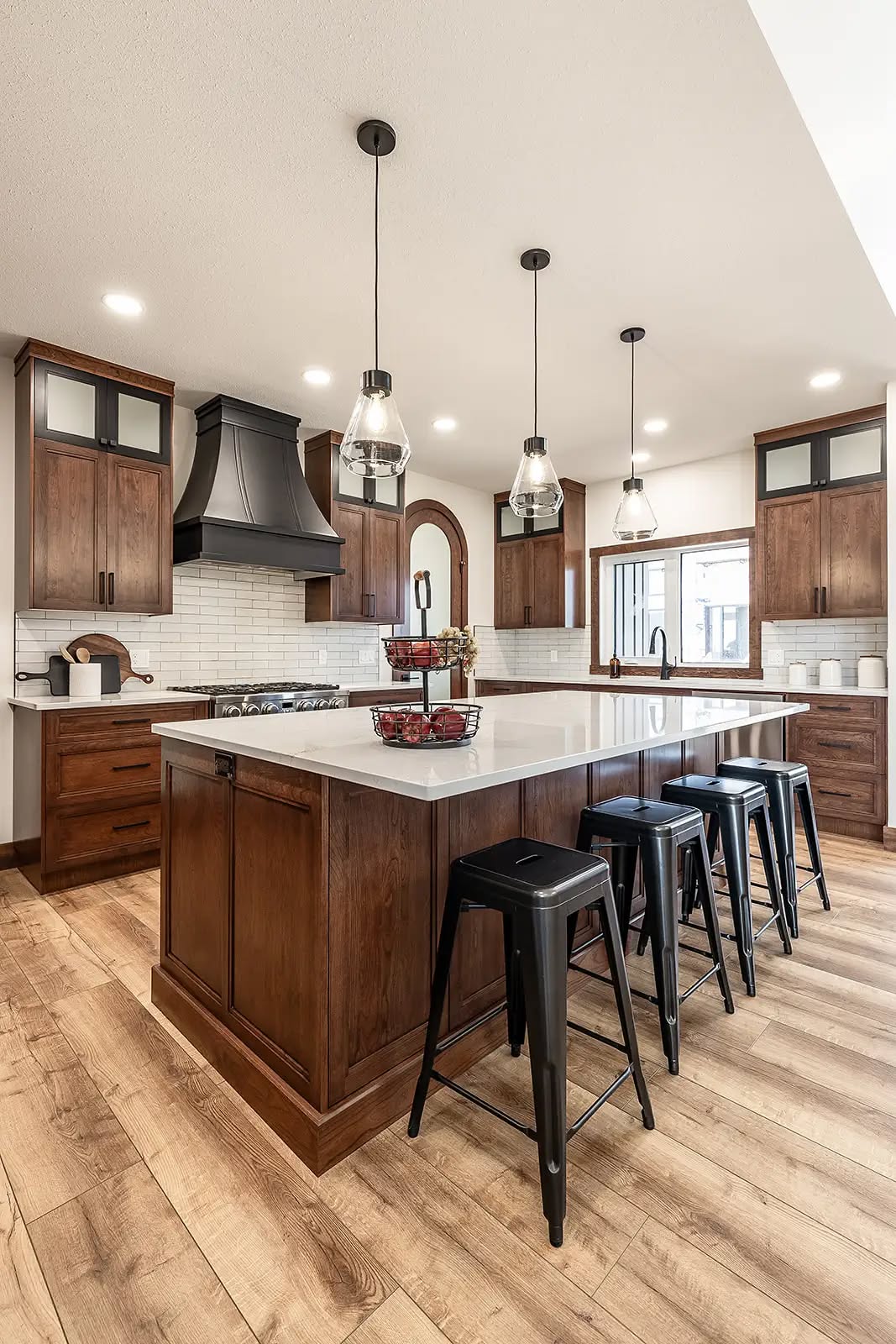 Spacious kitchen with wooden cabinets, white countertops, and a large island. Four black stools surround the island under modern pendant lights. Cozy and inviting.