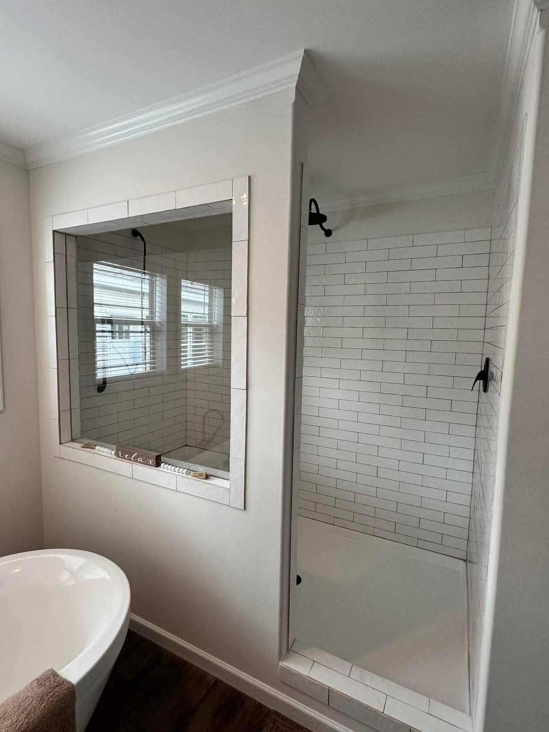 Modern bathroom with a glass shower enclosure and white subway tiles. A white bathtub sits nearby on a dark wood floor, creating a clean, serene ambiance.
