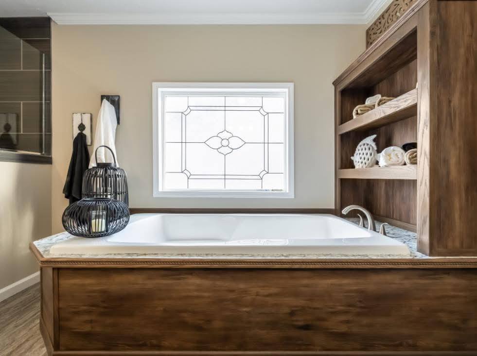 Elegant bathroom with a luxurious white bathtub set in a wooden frame. A stained glass window adds a vintage touch. Shelves with towels offer a cozy feel.