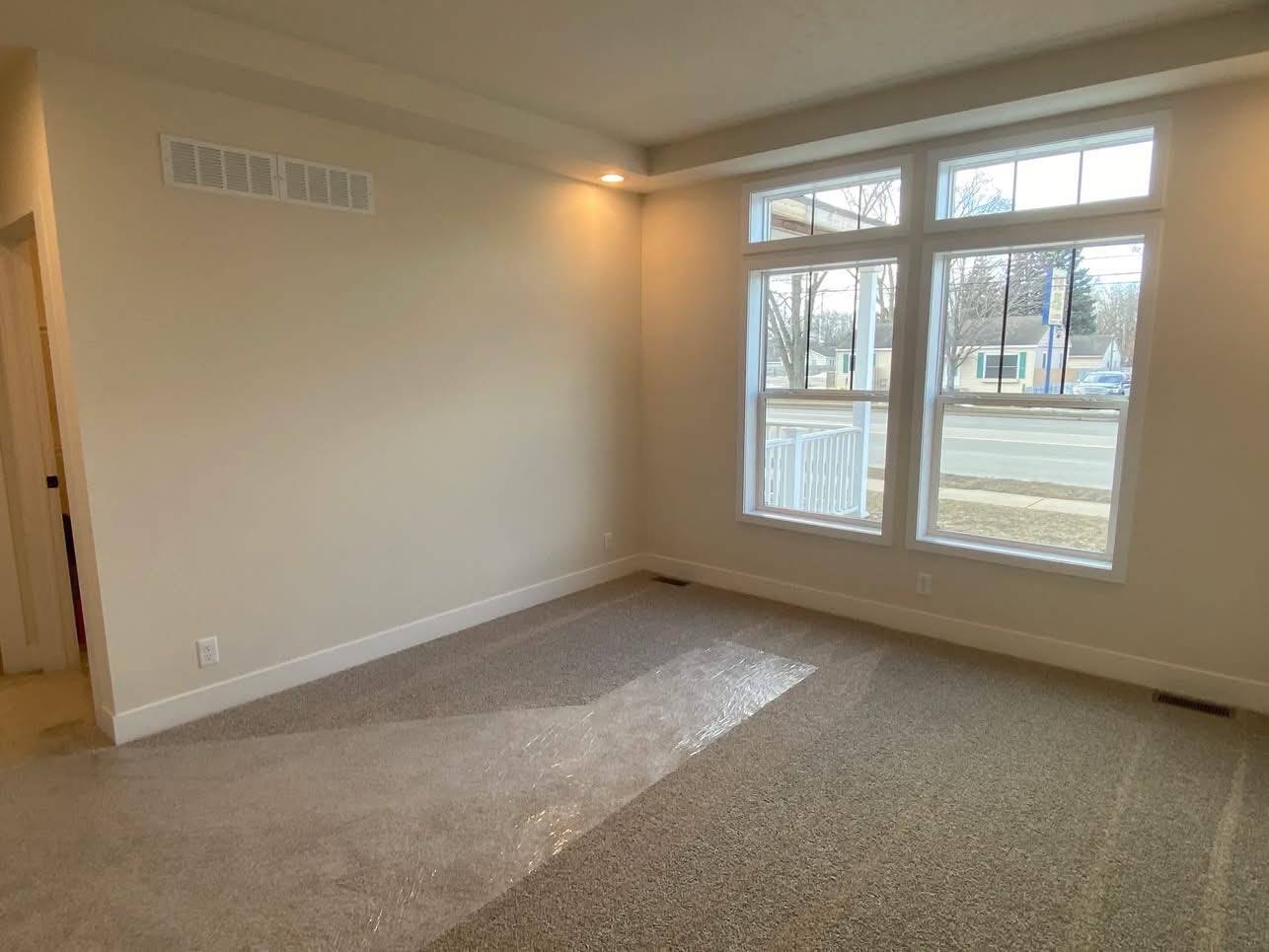 An empty room with beige walls and carpeted floor, featuring large windows overlooking a street with trees and houses. Soft lighting creates a calm atmosphere.
