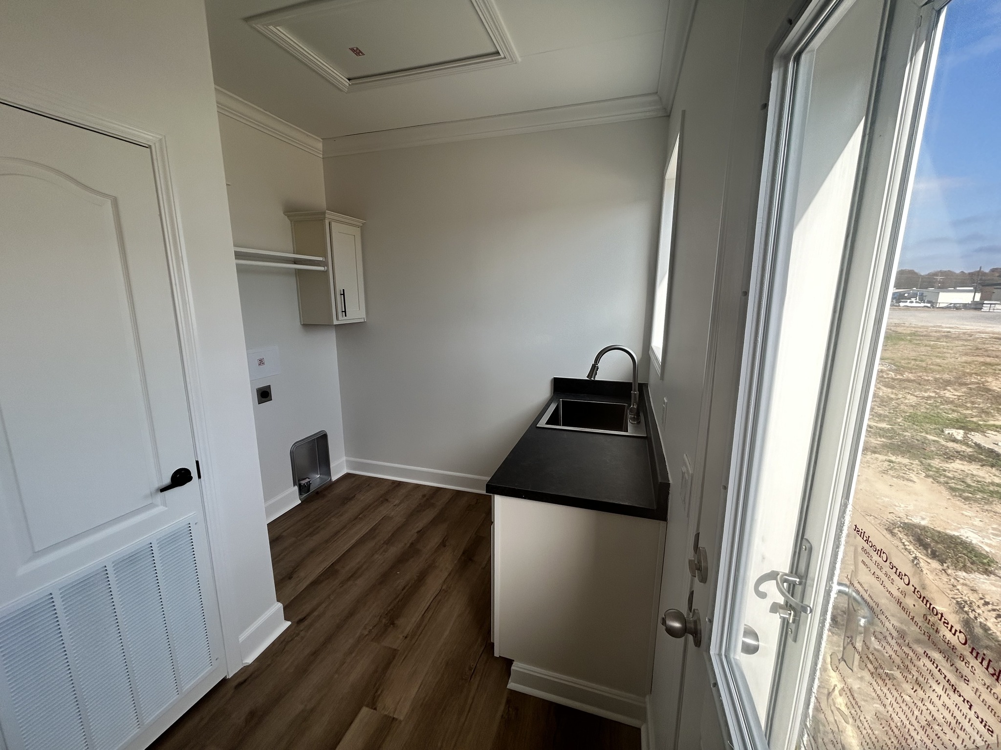 A small, sunlit utility room with light wood floors and white walls. It features a sleek, black countertop with a sink beneath a window.