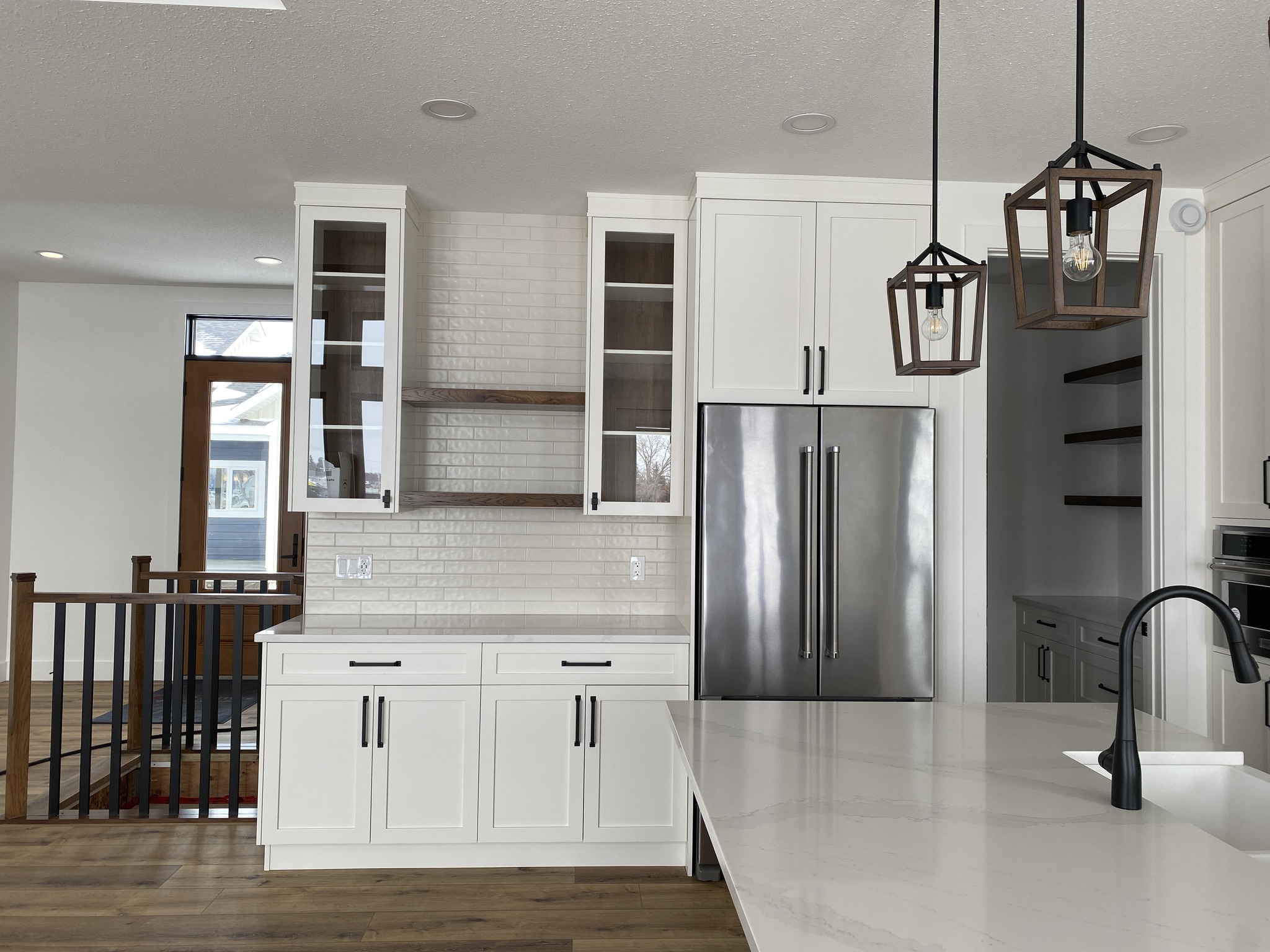 Modern kitchen with white cabinets, stainless steel fridge, and two geometric pendant lights over a sleek island. Wood accents and open shelving add warmth.
