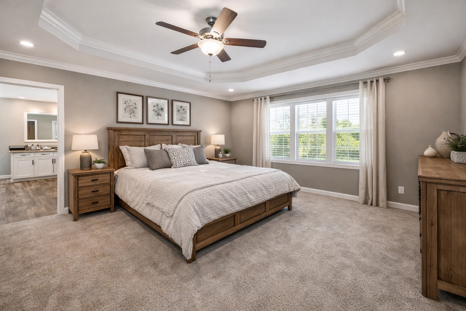 A cozy bedroom with a wood bed frame and beige bedding. Soft lighting emits from lamps on nightstands. A ceiling fan hangs above, and a large window with curtains allows natural light in. The room has a calm, inviting ambiance.