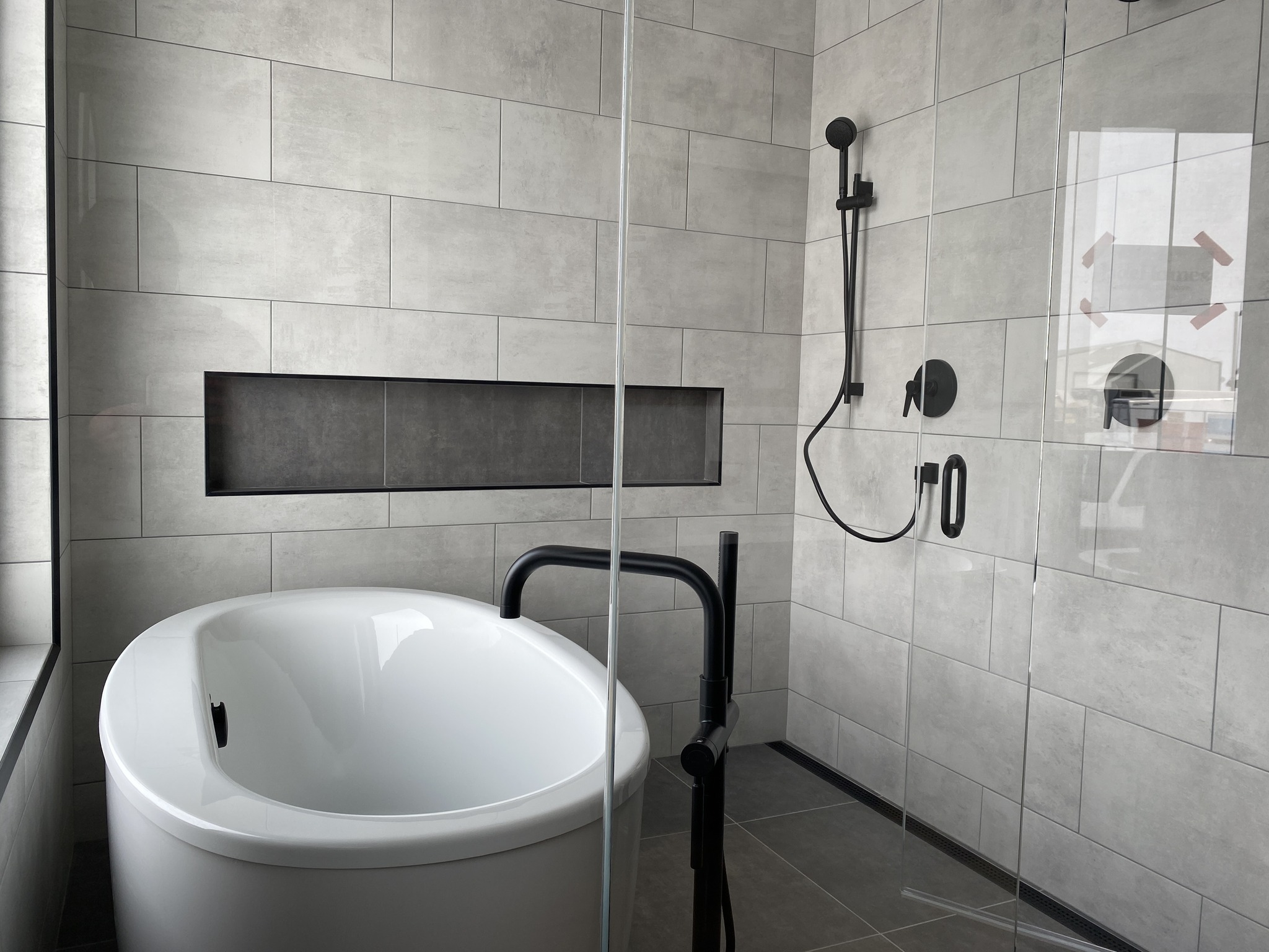 Modern bathroom with a freestanding white bathtub, matte black fixtures, and gray tiled walls. A glass shower enclosure adds a sleek, minimalist feel.