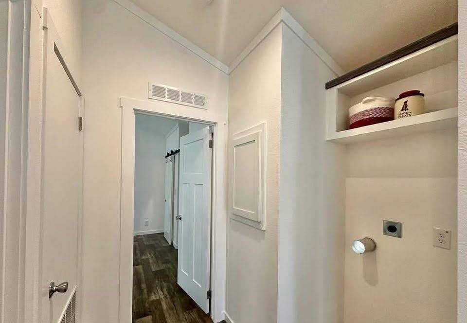 Narrow white hallway with a closed door at the end, open closet shelving on the right with decorative jars, and a wood floor creating a clean, simple look.
