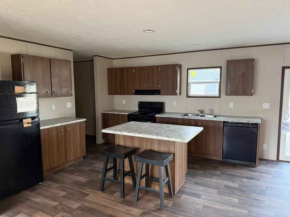 A modern kitchen with brown wooden cabinets, black appliances, and a marble-pattern countertop. Two black stools sit at the island, with a cozy, uncluttered feel.