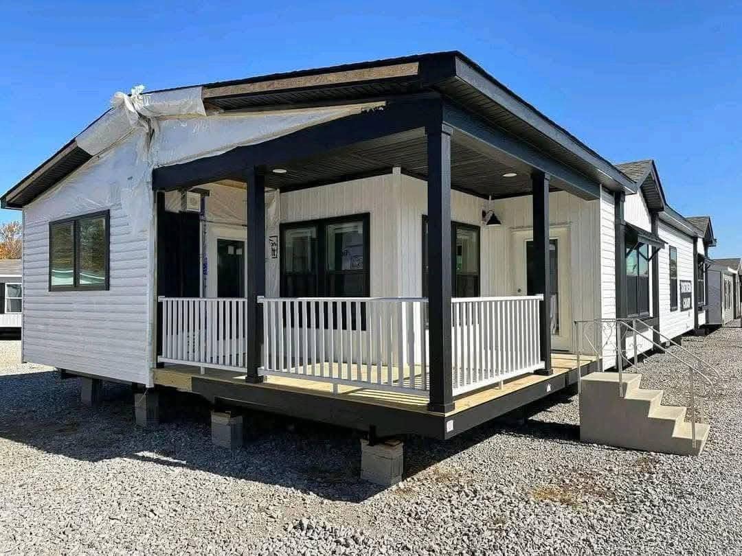 Modern modular home with black and white exterior, featuring a wraparound porch, white railing, and steps. Sunny day with gravel surroundings.