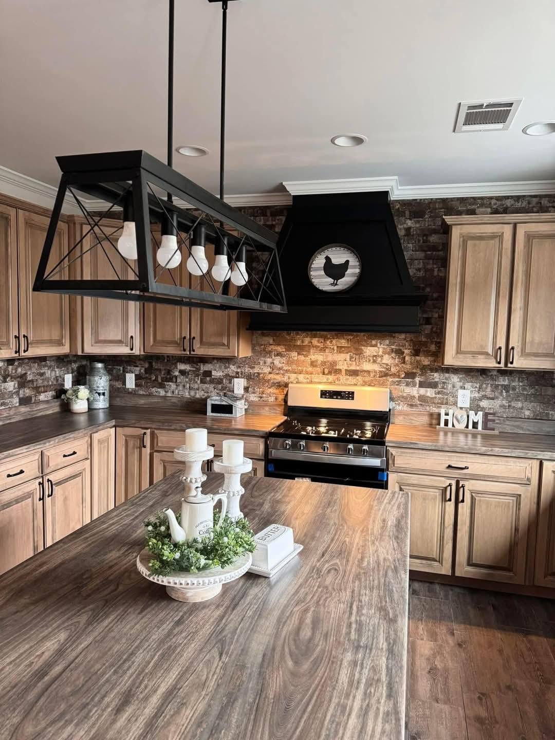 Rustic kitchen with wooden cabinets and brick backsplash. A large island features candles and greenery. Black chandelier above adds a modern touch.