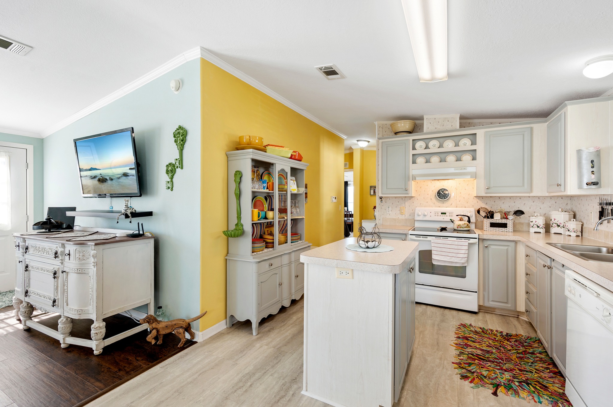 Bright kitchen and living area with light blue and yellow walls. White cabinets and kitchen island; a colorful rug and plates add vibrancy. Cozy and inviting atmosphere.