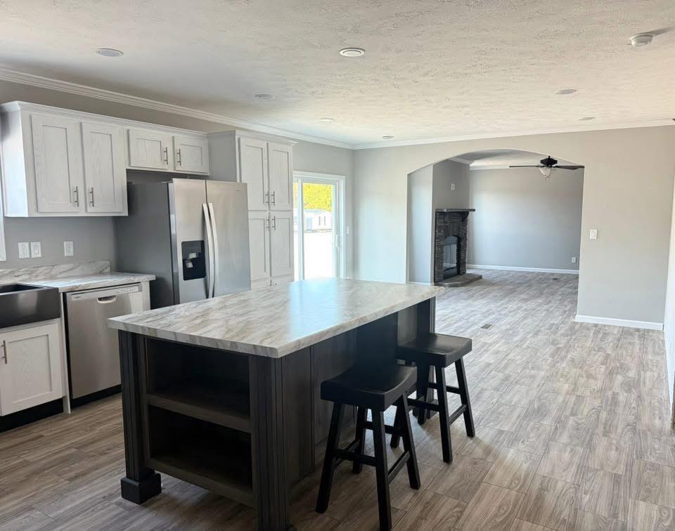 Modern kitchen with light wood cabinets, stainless steel appliances, and a marble island with two stools. Open living area with wood flooring.