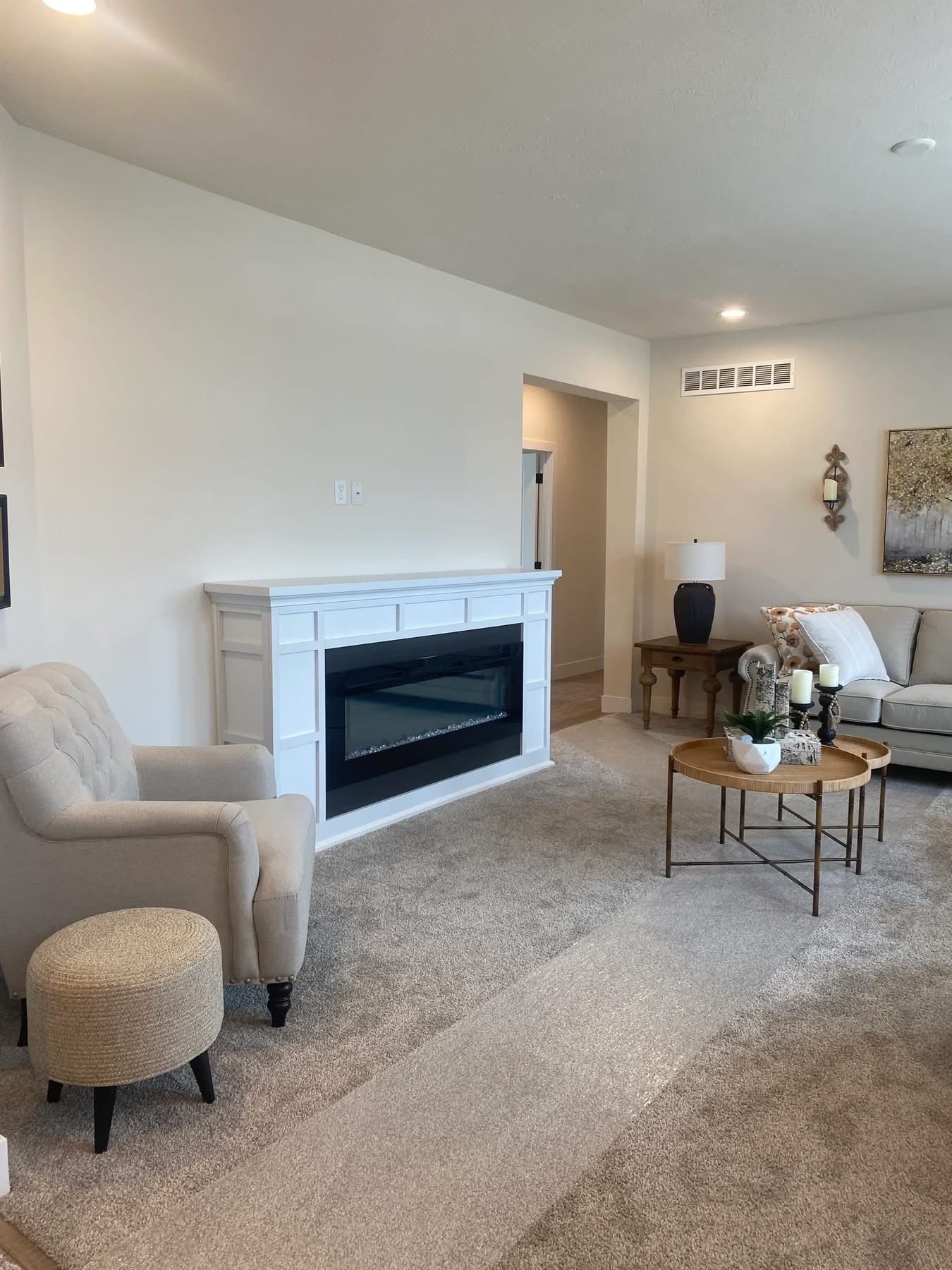 Stylish living room with a white electric fireplace, beige armchair, and round coffee table. Neutral tones and soft lighting create a cozy atmosphere.