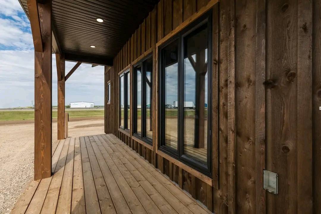 Wooden porch with dark-stained siding, large windows, and a black ceiling. It overlooks a rural landscape with a cloudy sky and distant buildings.