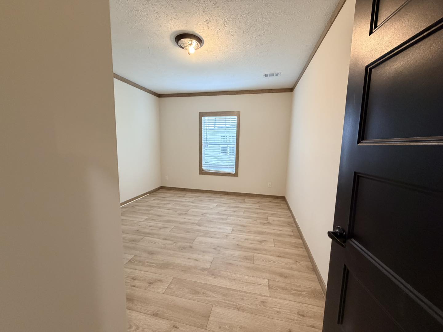 Empty room with light wood flooring, cream walls, and a dark ceiling light fixture. A single window with blinds is centered on the far wall.
