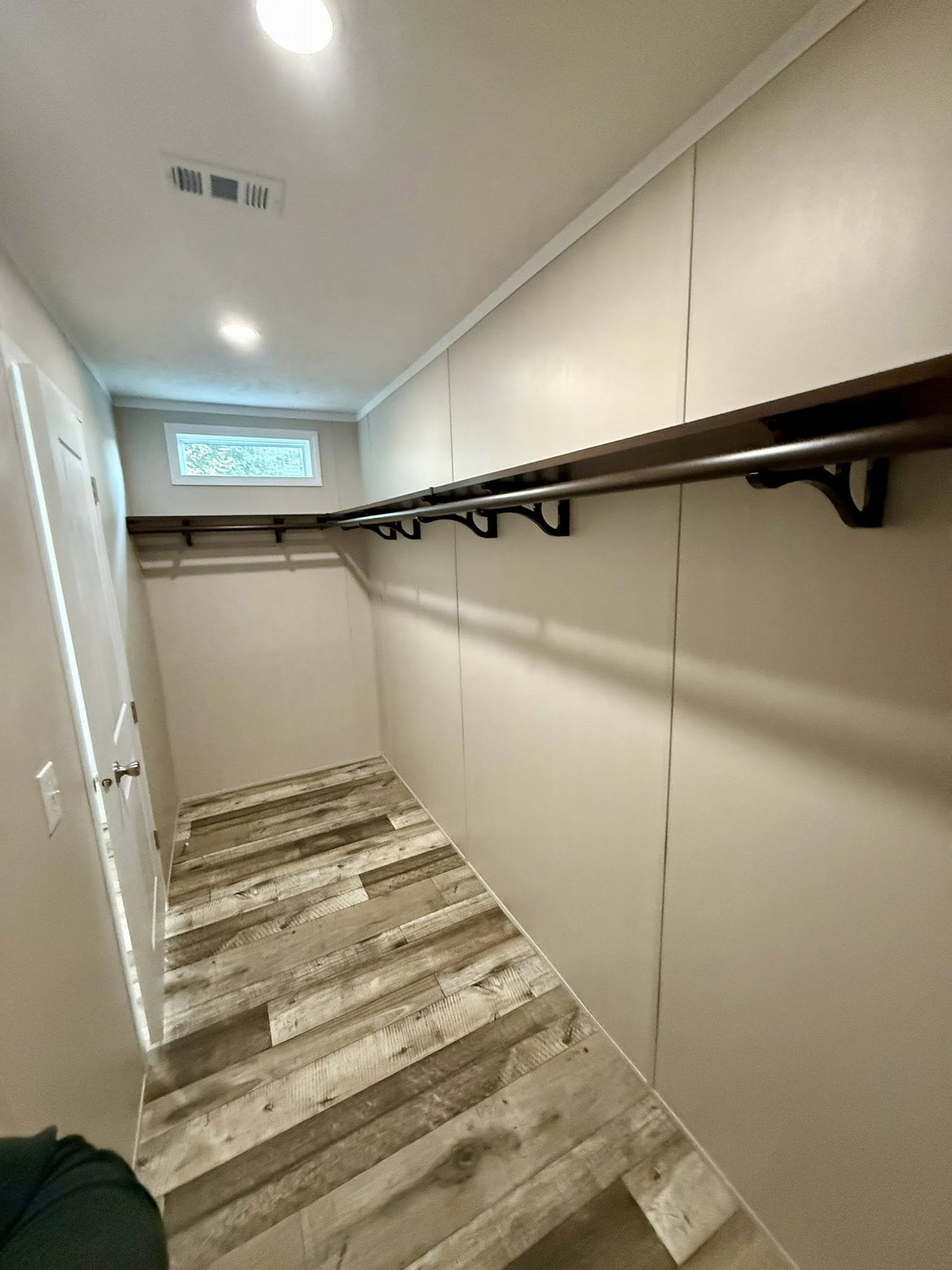 Narrow walk-in closet with beige walls, a long wooden clothes rail, and rustic wood floorboards. A small window lets in natural light, creating a cozy feel.