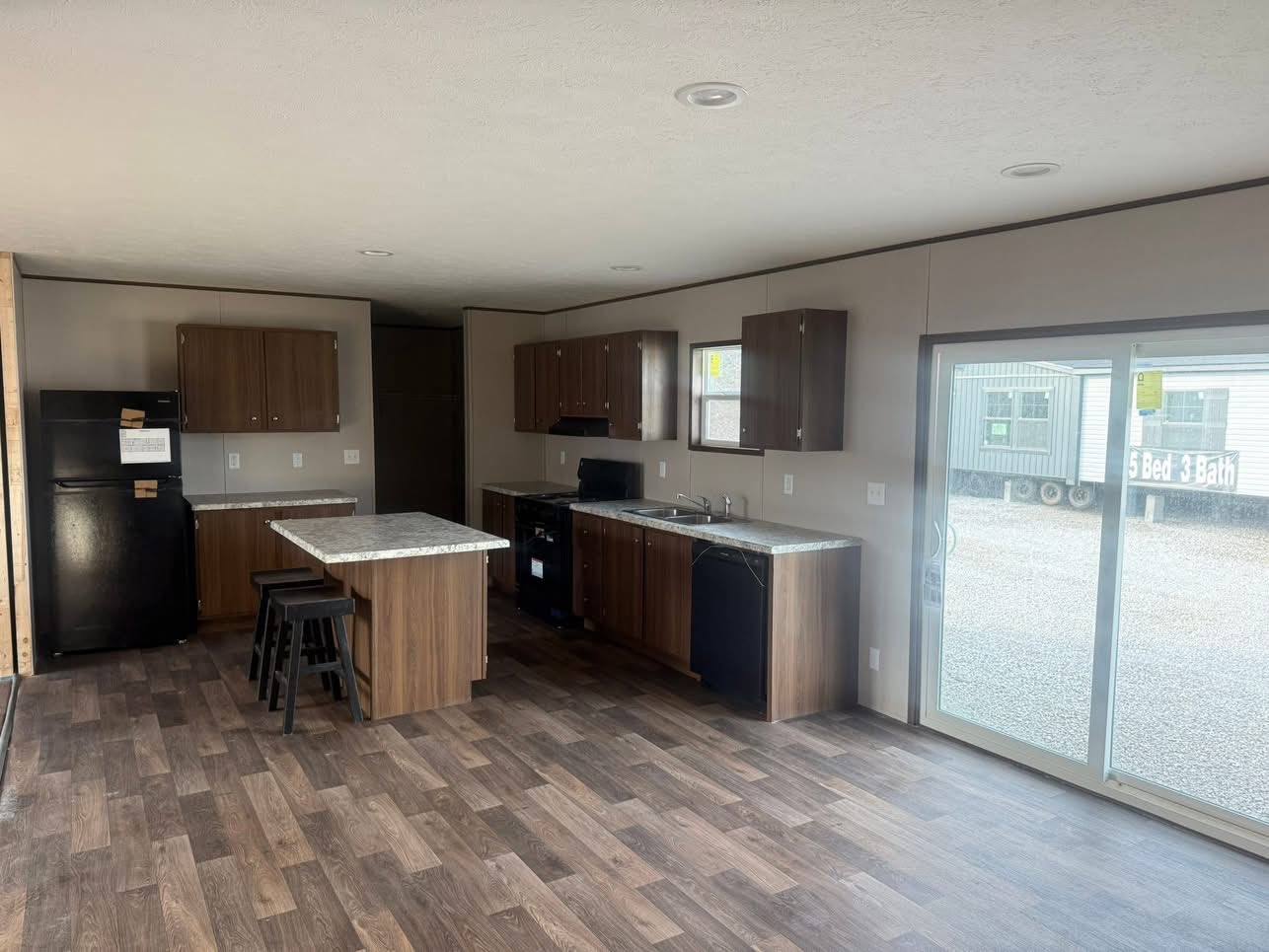 Spacious kitchen in a mobile home with wood cabinets, black appliances, and an island. Large sliding glass doors provide natural light, creating a cozy atmosphere.