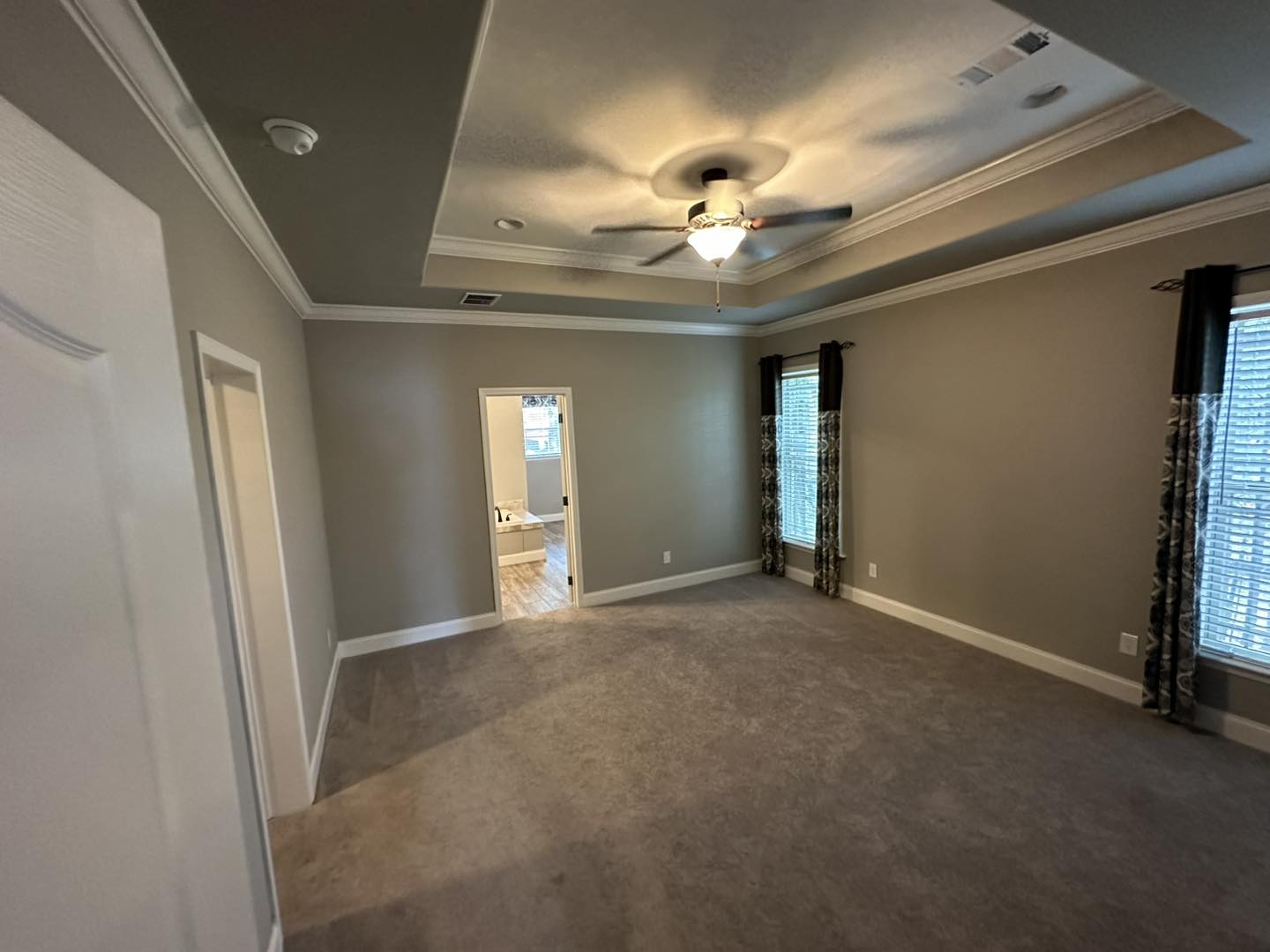 Spacious bedroom with a tray ceiling and ceiling fan, featuring large windows with dark curtains. A doorway leads to a bright en-suite bathroom.