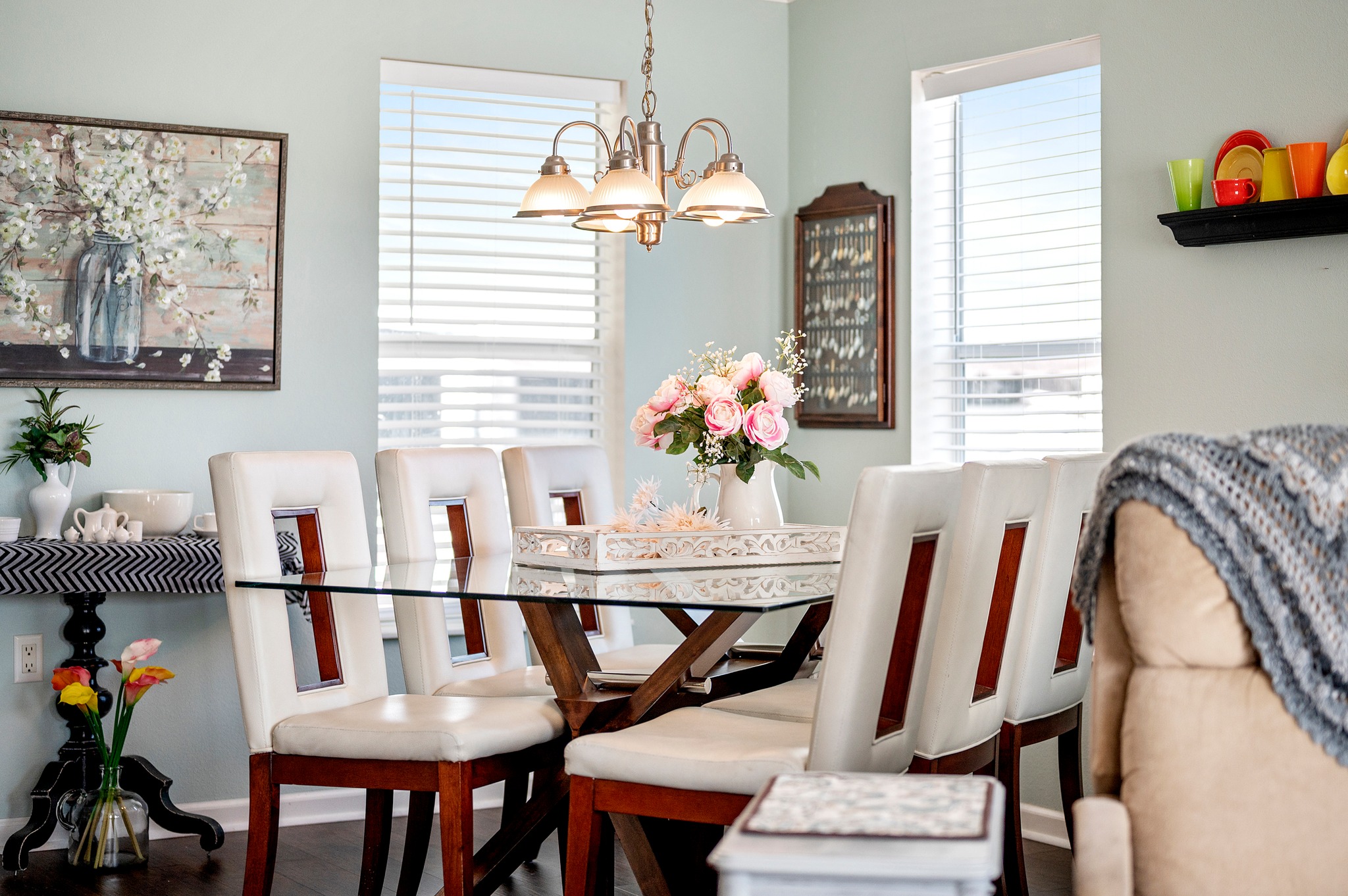 A cozy dining room with a glass-topped table and eight white chairs. A chandelier hangs above, with a bouquet of pink flowers as the table centerpiece. Warm light filters through the windows.