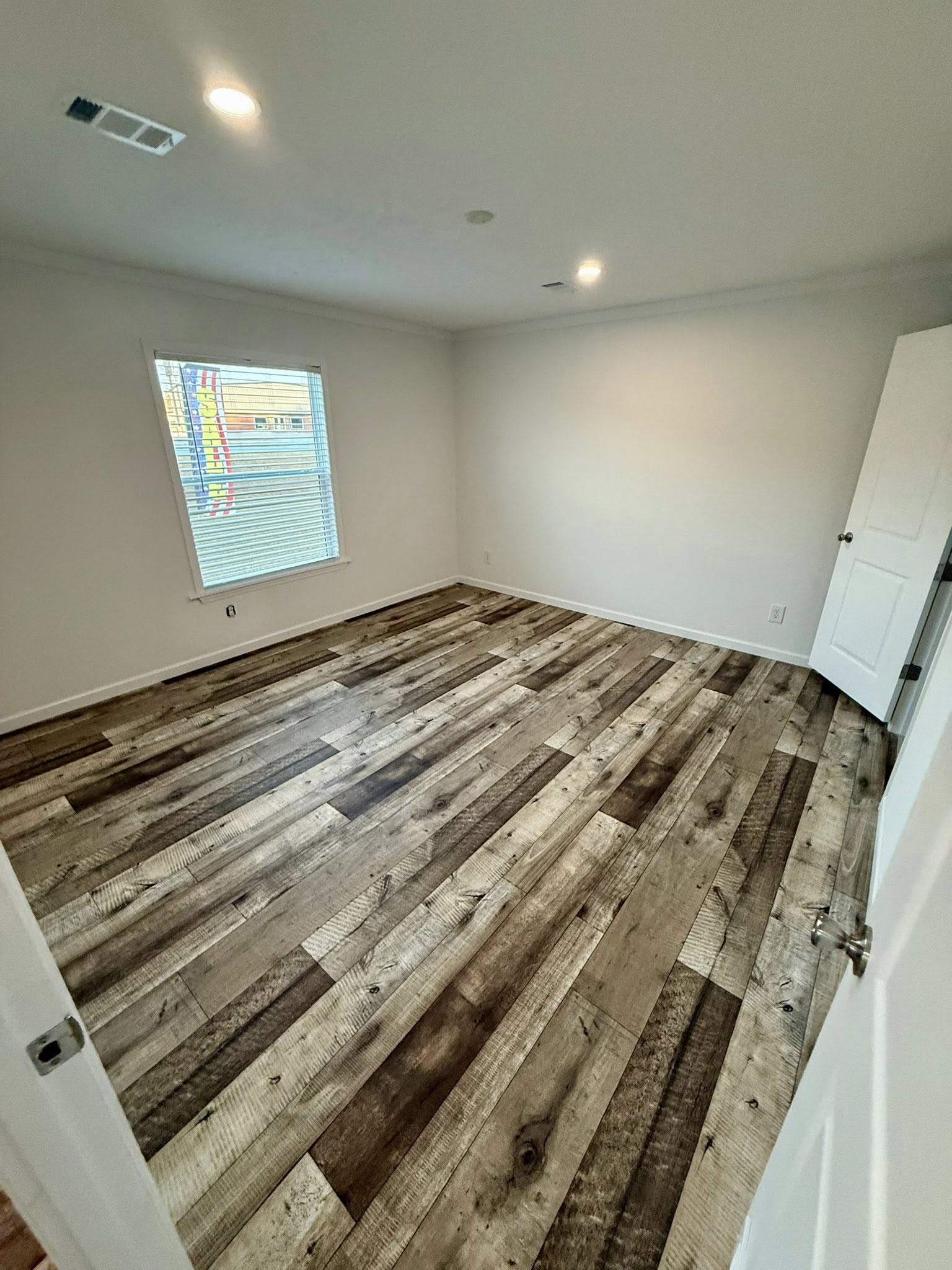 Empty room with rustic, wide-plank wooden flooring and white walls. A single window on the left lets in soft light, and a door is ajar on the right.