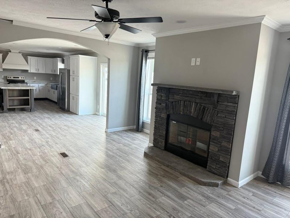 Spacious open-plan room with light wood flooring; features include a ceiling fan, stone fireplace, and a kitchen with white cabinets and stainless appliances.