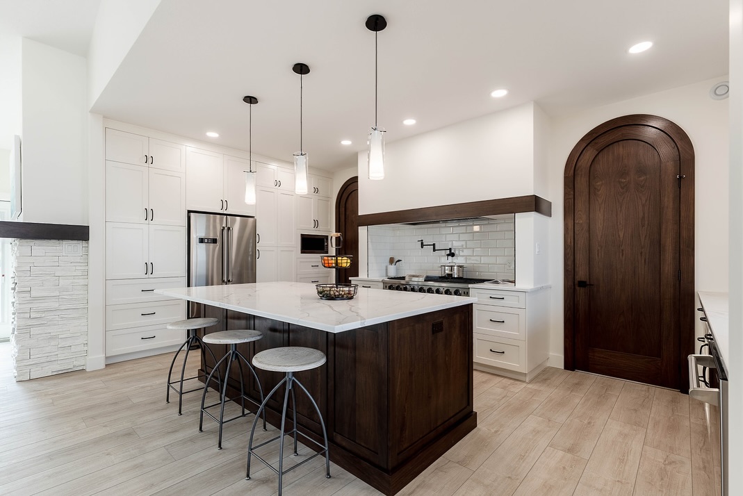 Modern kitchen with a large marble island, dark wood base, and four stools. White cabinets, stainless steel fridge, arched wooden door, and pendant lights create a sleek, inviting space.