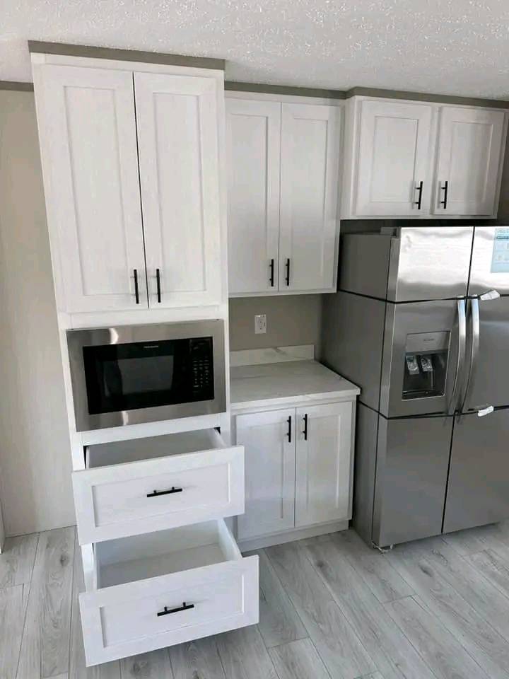 Modern kitchen featuring white cabinets with black handles, a built-in microwave, open drawers, and a stainless steel refrigerator against light wood flooring.