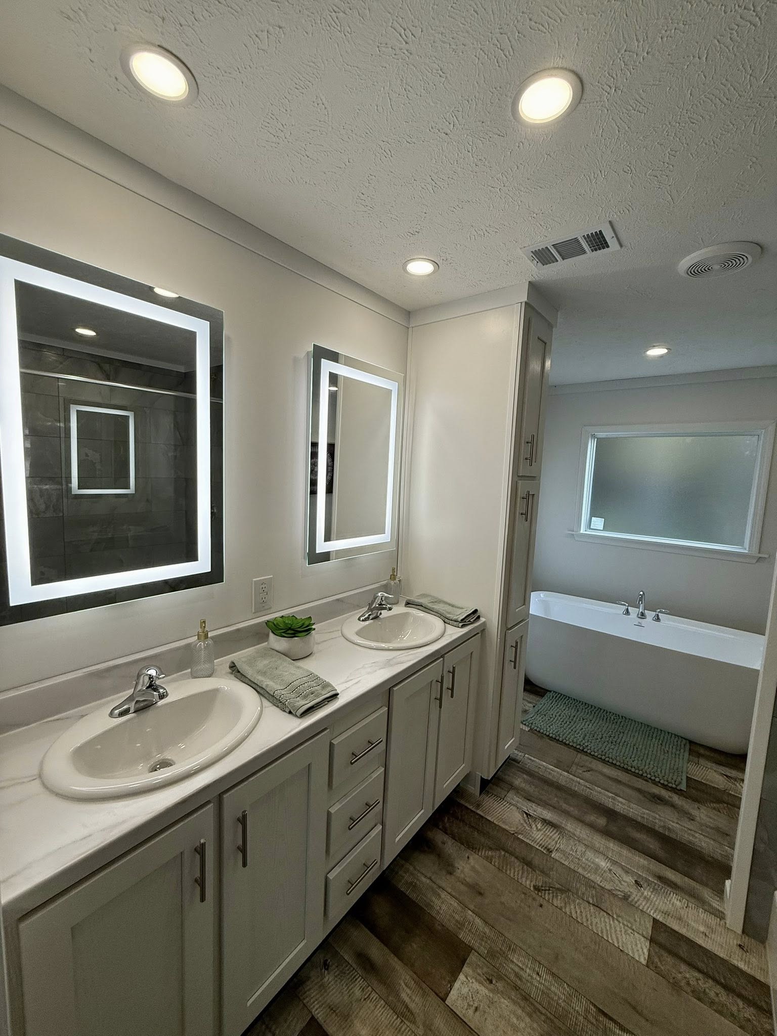 Modern bathroom with dual sinks, backlit mirrors, and a freestanding white bathtub. Wooden floor enhances warmth; overall clean and relaxing ambiance.