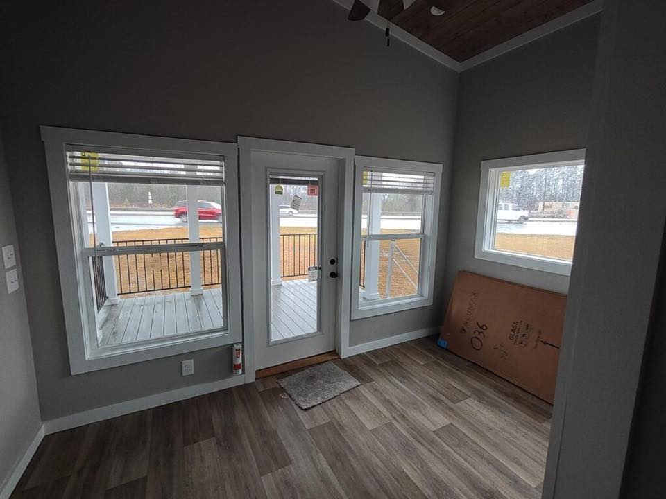A small, unfurnished room with gray walls and wood flooring features a white-framed glass door and windows overlooking a porch and road, creating a calm atmosphere.
