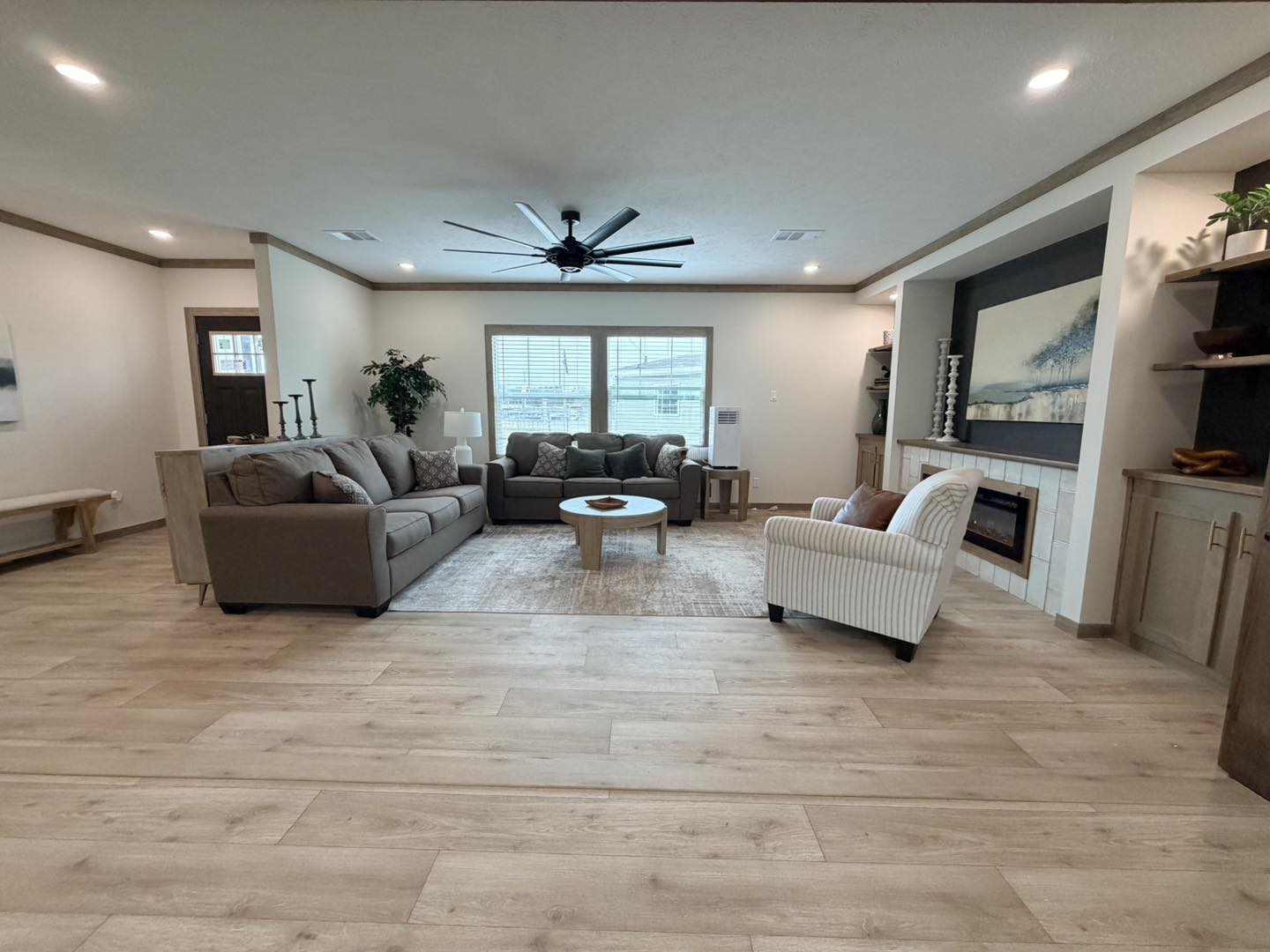 Spacious living room with wooden floors, gray sectional sofa, and striped armchair. Features a round coffee table, large window, and modern fireplace. Cozy and inviting.
