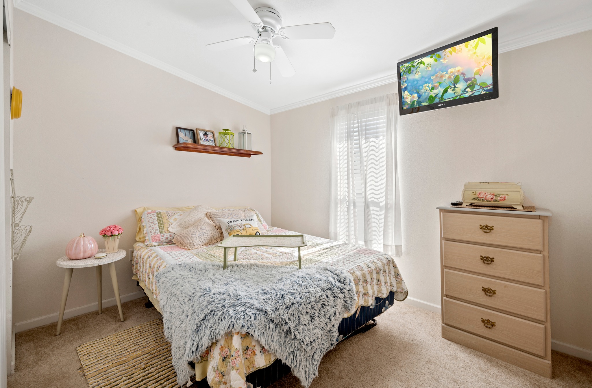 Cozy bedroom with beige walls, a bed with floral and fuzzy blankets, a TV mounted high, a small table with decorations, and soft, natural light filtering through sheer curtains.
