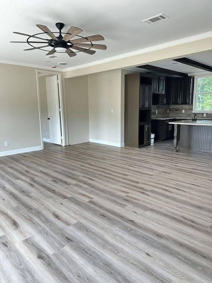 Modern room with light wood flooring, a ceiling fan, neutral walls, and an open kitchen featuring dark cabinets and a granite countertop.