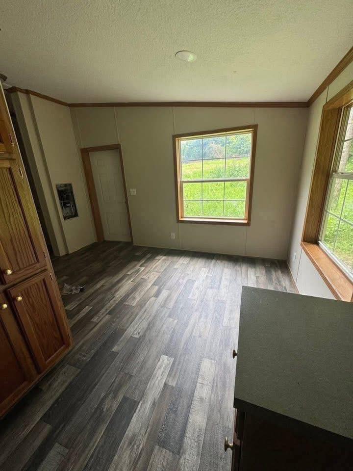 A room with dark wood flooring, beige walls, and large windows framing a green outdoor view. The space has a warm, inviting feel with wood cabinets.