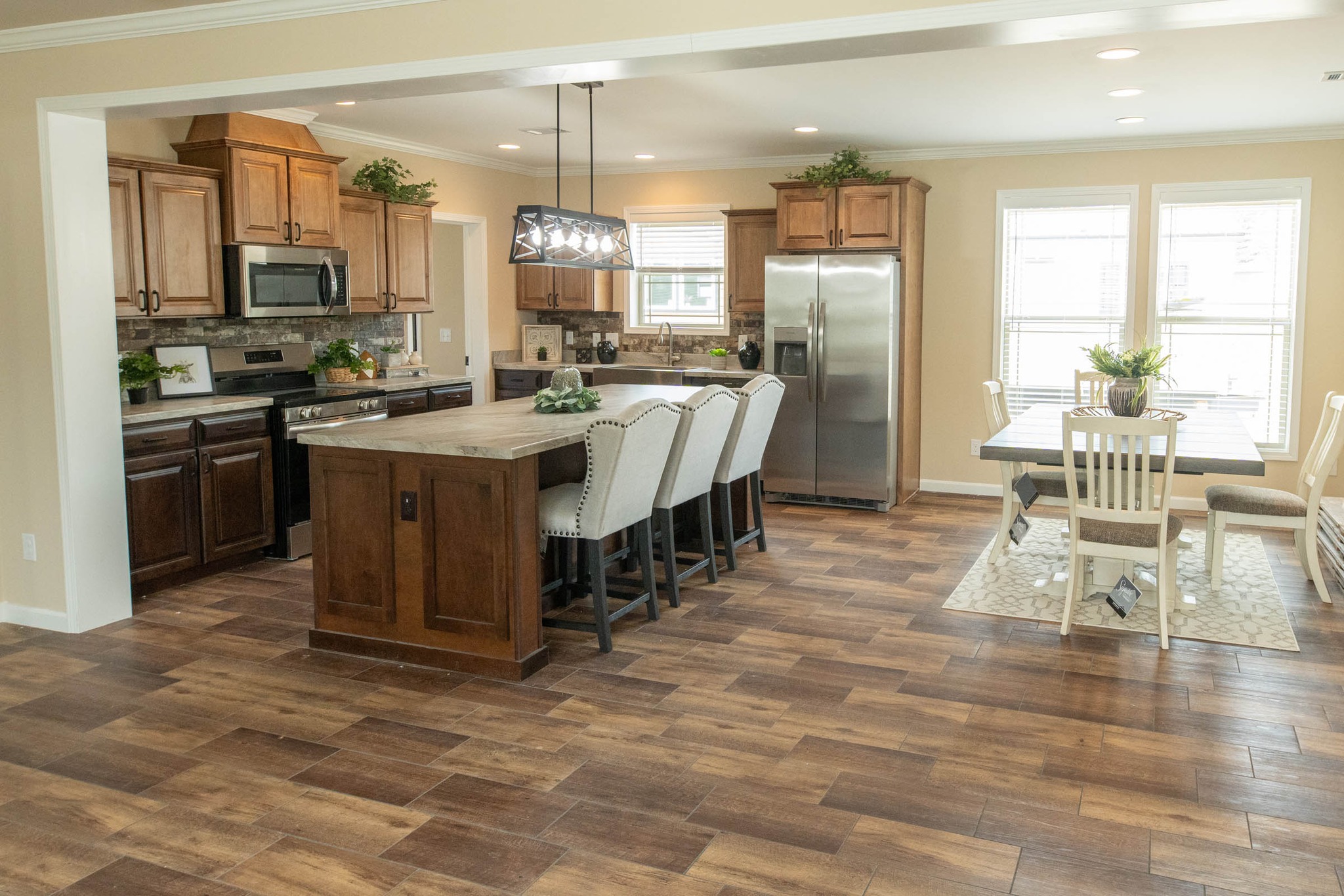 A spacious kitchen with wooden cabinets, stainless steel appliances, and a large island with chairs. A dining table is set near two large windows.