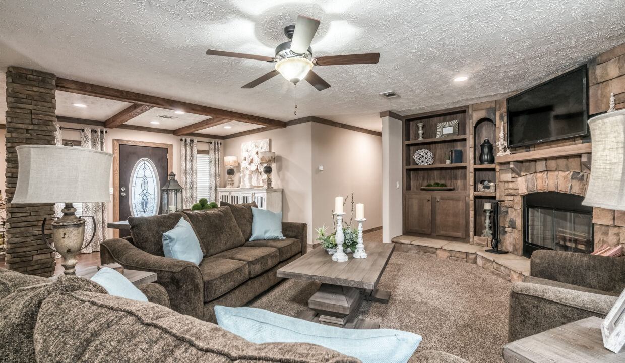 Cozy living room with a ceiling fan, brown sofas, and a wooden coffee table. Features a stone fireplace, built-in shelves, and soft lighting. Warm, welcoming ambiance.