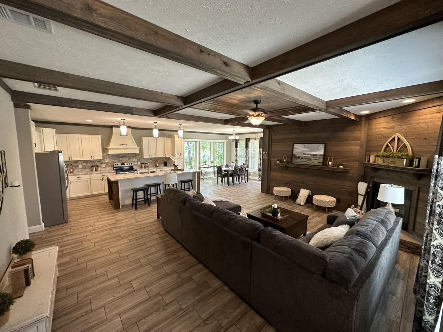 Open-concept living space with wooden beams and earthy tones. Features a kitchen island, cozy sectional sofa, decorative fireplace, and dining area. Warm and inviting ambiance.