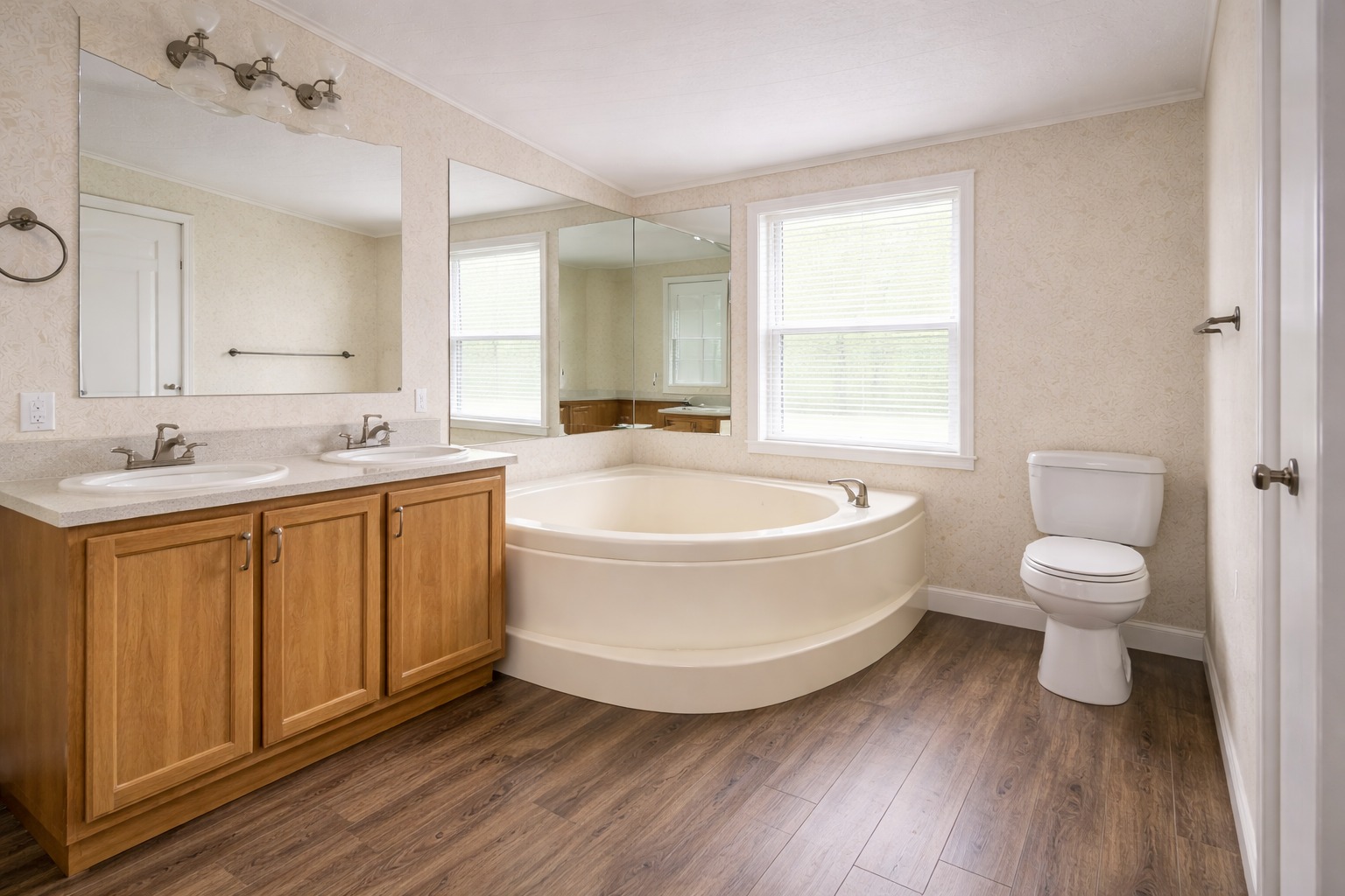 Spacious bathroom with light wood flooring, dual sinks in a wood vanity, large mirrors, a corner bathtub, and a toilet. Bright and clean ambiance.