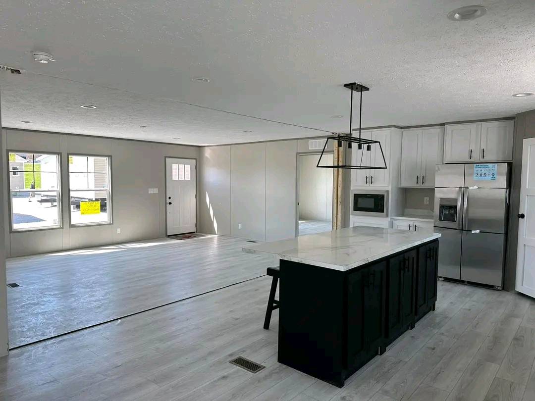 Open-concept kitchen and living room with bright natural light. Features light wood flooring, an island with a modern light fixture, and stainless steel appliances. The space is minimal and inviting.