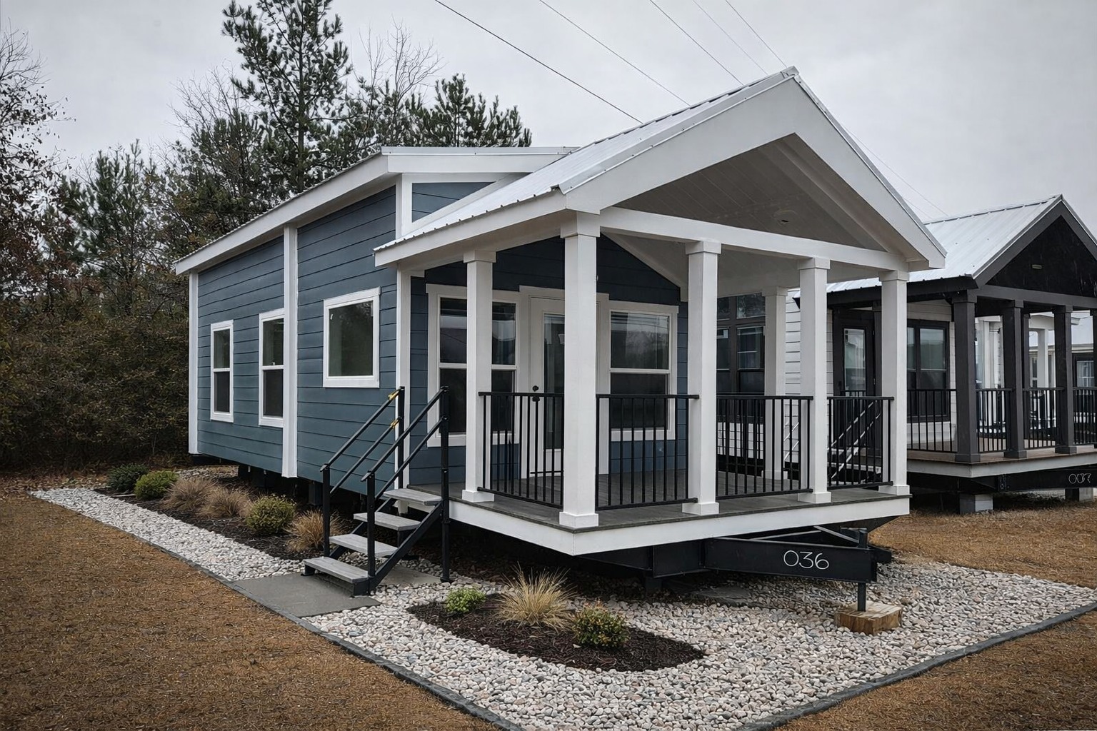 A small, modern tiny house with a blue exterior, white trim, and a covered front porch. It's set on a gravel path, surrounded by sparse shrubs, under an overcast sky.